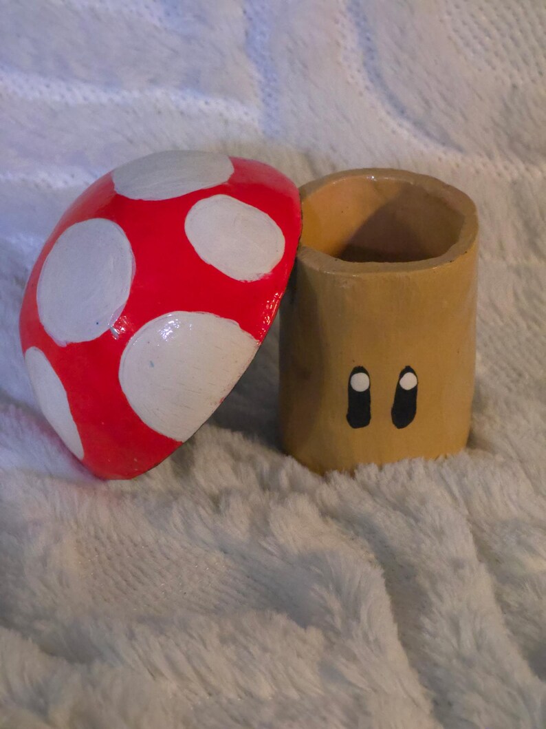 May include: A cylindrical tan container with two black and white oval eyes painted on the front, accompanied by a separate red mushroom cap lid featuring white circular spots. The object is constructed with a hollow interior and a smooth finish.