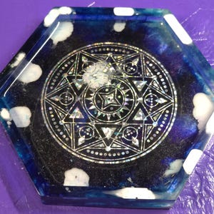 May include: A hexagonal resin coaster featuring a deep blue and black swirling background with white cloud-like accents. The center displays a detailed, silver-toned geometric star pattern with intricate lines and holographic elements.