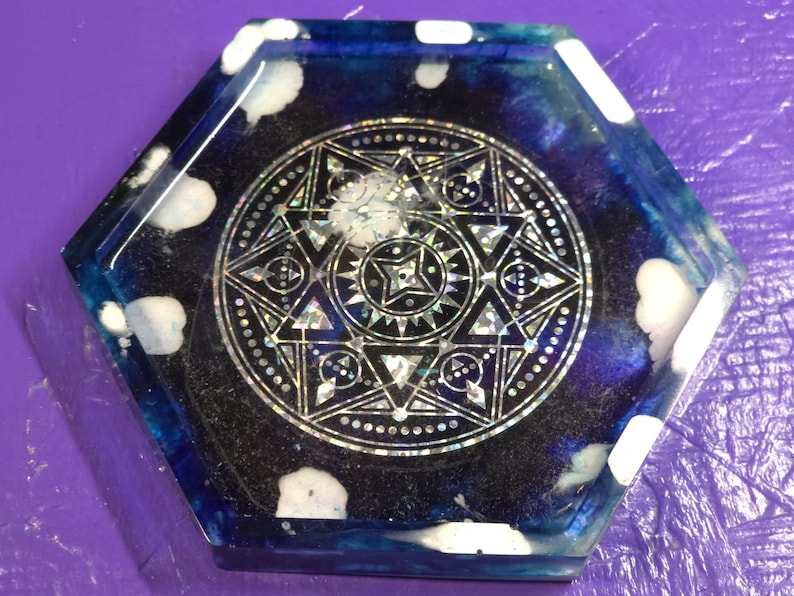 May include: A hexagonal resin coaster featuring a deep blue and black swirling background with white cloud-like accents. The center displays a detailed, silver-toned geometric star pattern with intricate lines and holographic elements.