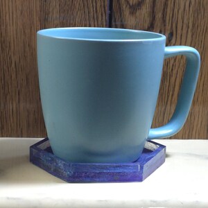 Pode incluir: Uma caneca de cer&acirc;mica azul-clara com acabamento mate suave e pega curva, assente num porta-copos hexagonal que apresenta um padr&atilde;o de resina marmoreada em azul e roxo profundo com uma textura transl&uacute;cida e brilhante nas bordas.