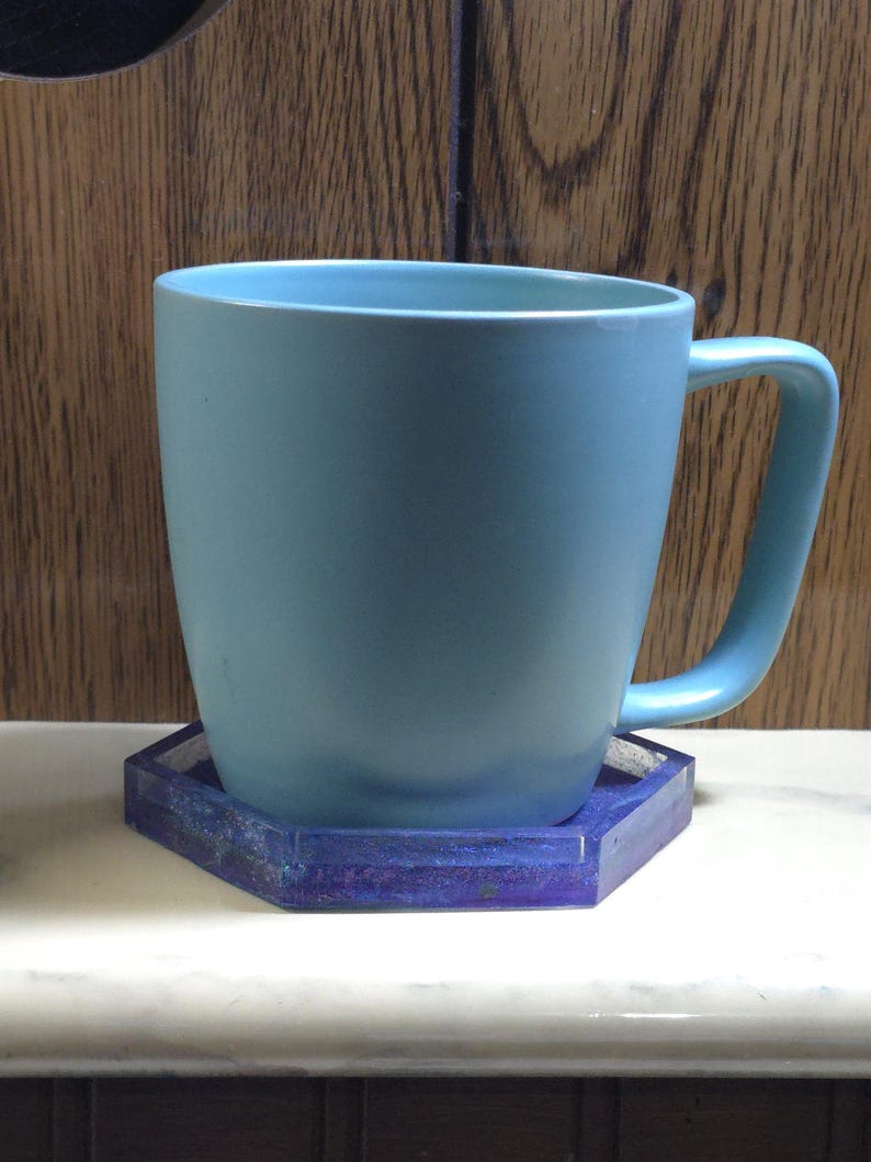Pode incluir: Uma caneca de cer&acirc;mica azul-clara com acabamento mate suave e pega curva, assente num porta-copos hexagonal que apresenta um padr&atilde;o de resina marmoreada em azul e roxo profundo com uma textura transl&uacute;cida e brilhante nas bordas.