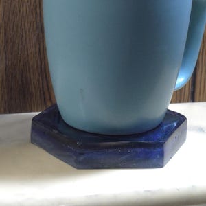 May include: A light blue ceramic mug sits on top of a dark blue hexagonal coaster. The mug has a smooth, matte finish and a curved handle on the right side. The coaster features a deep, glossy, dark blue surface with subtle variations in tone.