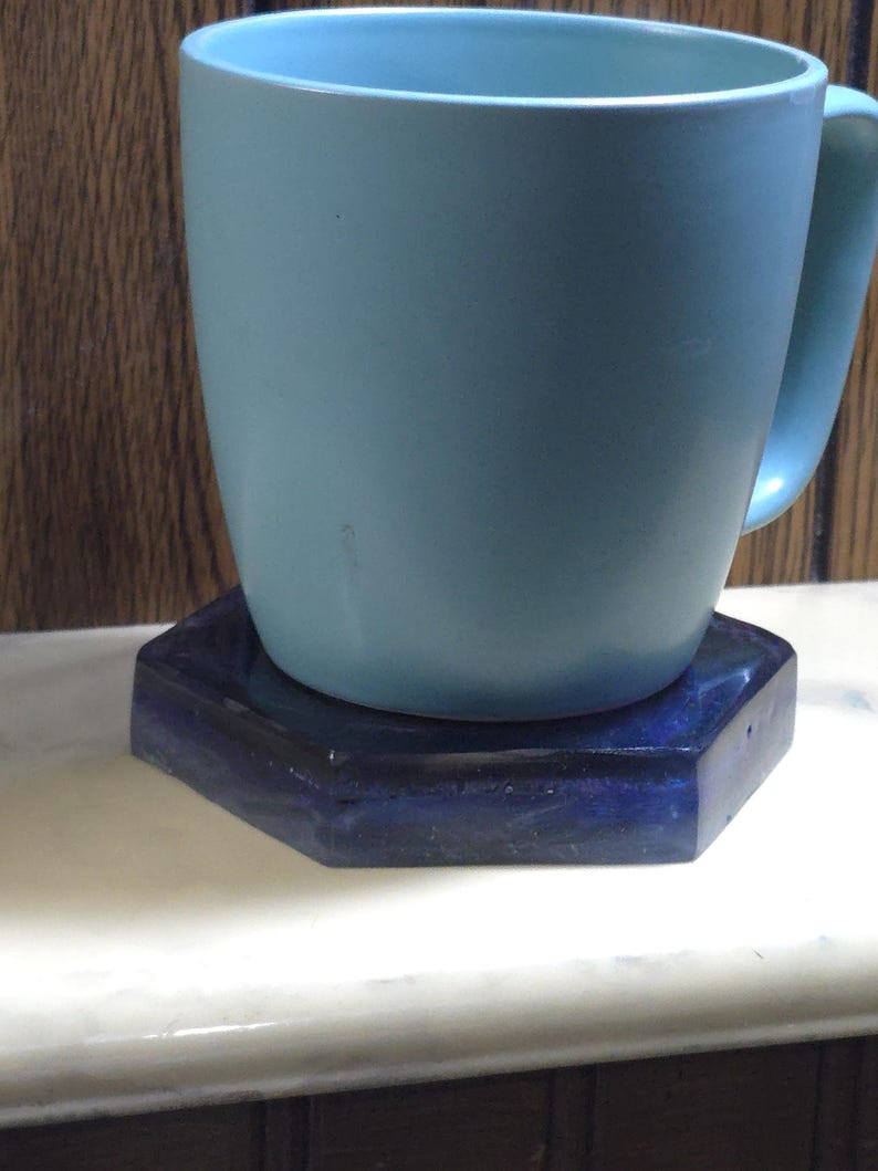 May include: A light blue ceramic mug sits on top of a dark blue hexagonal coaster. The mug has a smooth, matte finish and a curved handle on the right side. The coaster features a deep, glossy, dark blue surface with subtle variations in tone.