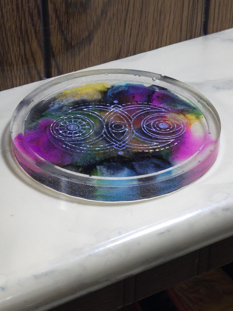 May include: A circular clear resin coaster featuring a swirling multi-colored interior with shades of magenta, blue, yellow, and black. The surface is embossed with a white geometric design consisting of three interconnected circular motifs.