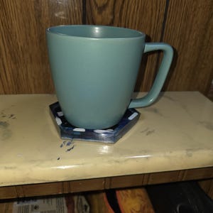 May include: A matte sage green ceramic mug with a rounded body and a C-shaped handle, resting on a hexagonal coaster featuring a dark blue and black marbled pattern with small white accents along the edges of the coaster's surface.