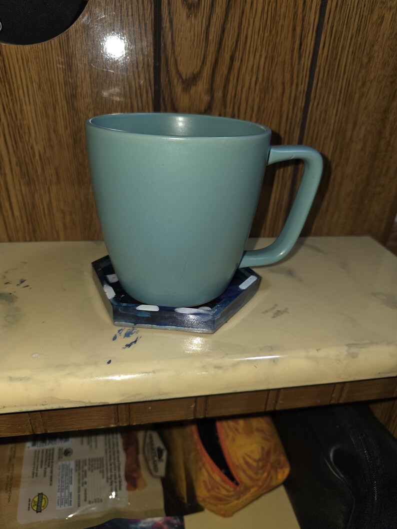 May include: A matte sage green ceramic mug with a rounded body and a C-shaped handle, resting on a hexagonal coaster featuring a dark blue and black marbled pattern with small white accents along the edges of the coaster's surface.