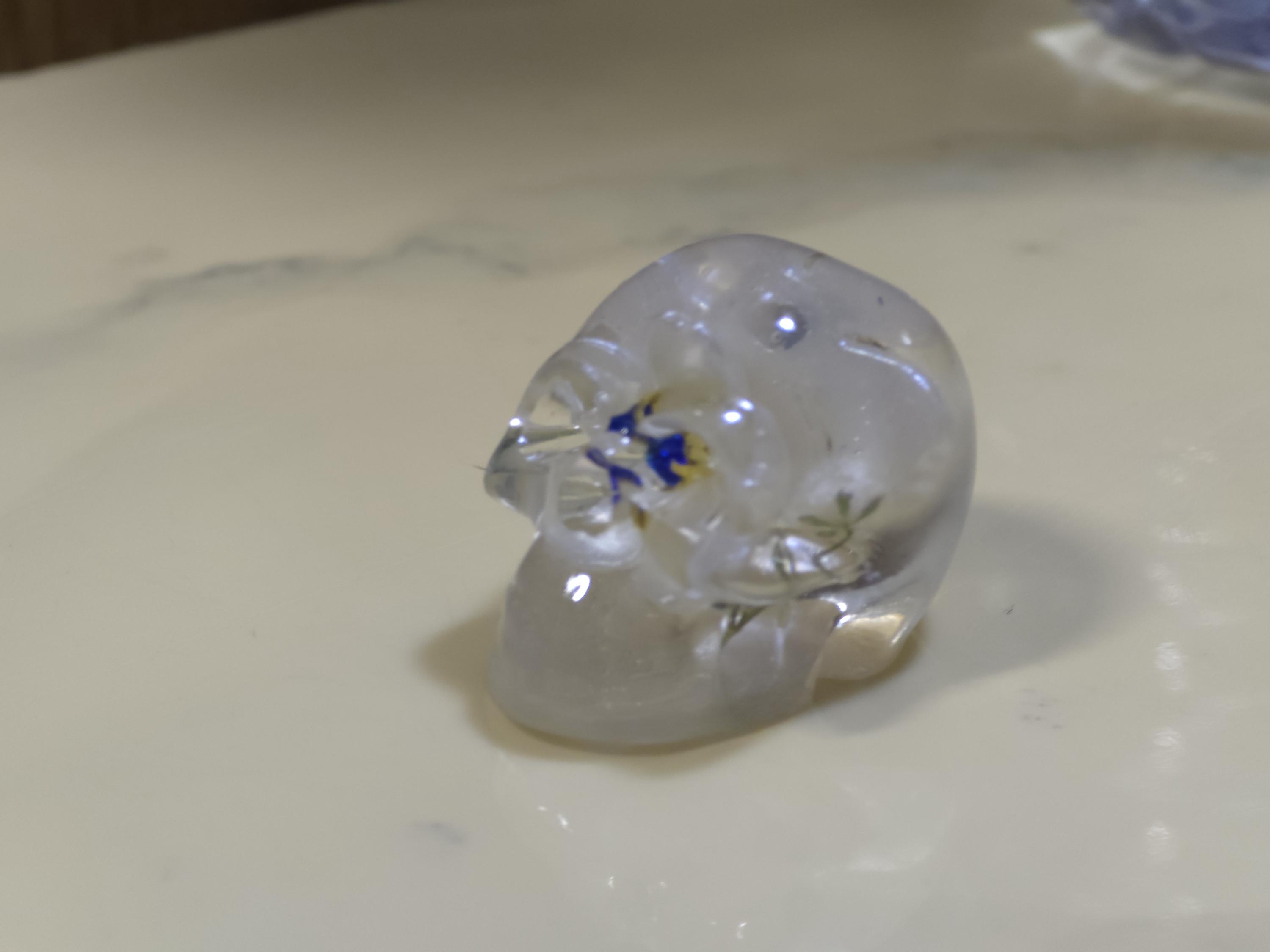 May include: A small, clear, translucent skull figurine crafted from resin. The skull features embedded decorative elements, including small blue and yellow floral accents visible within the transparent material, creating a detailed and reflective surface.