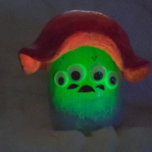 May include: A glowing green creature figurine with four white eyes and a dark mouth, topped with a large, curved, reddish-orange mushroom-shaped cap. The body emits a bright light, highlighting the facial features against the dark, textured surface.