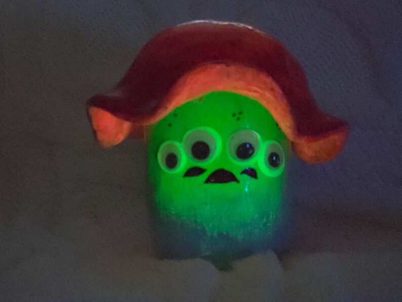 May include: A glowing green creature figurine with four white eyes and a dark mouth, topped with a large, curved, reddish-orange mushroom-shaped cap. The body emits a bright light, highlighting the facial features against the dark, textured surface.
