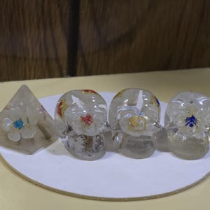 May include: A clear resin pyramid and three clear resin skulls, each containing a white flower with a colored center. The pyramid flower has a blue center, while the skulls feature flowers with red, yellow, and blue centers respectively.