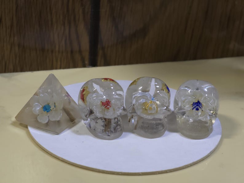 May include: A clear resin pyramid and three clear resin skulls, each containing a white flower with a colored center. The pyramid flower has a blue center, while the skulls feature flowers with red, yellow, and blue centers respectively.