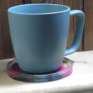 May include: A light blue ceramic mug with a smooth matte finish and a curved handle, resting on a circular, translucent coaster featuring a vibrant, multi-colored swirl pattern of pink, blue, and yellow hues embedded within the clear material.