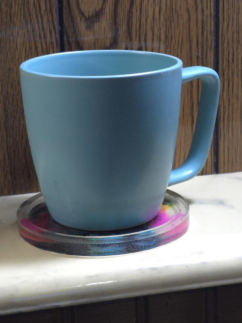 May include: A light blue ceramic mug with a smooth matte finish and a curved handle, resting on a circular, translucent coaster featuring a vibrant, multi-colored swirl pattern of pink, blue, and yellow hues embedded within the clear material.