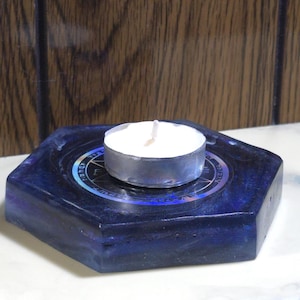 May include: A hexagonal dark blue resin candle holder with a smooth, glossy finish. Centered on the top surface is a circular metallic sticker featuring a black pentagram design surrounded by intricate, illegible text. A white tea light candle sits.