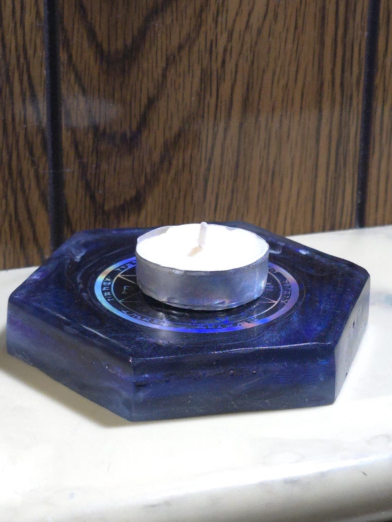 May include: A hexagonal dark blue resin candle holder with a smooth, glossy finish. Centered on the top surface is a circular metallic sticker featuring a black pentagram design surrounded by intricate, illegible text. A white tea light candle sits.