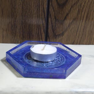 Pode incluir: Uma bandeja hexagonal de resina azul com um design circular central apresentando padr&otilde;es geom&eacute;tricos intrincados e motivos de estrelas. Uma pequena vela branca redonda com um inv&oacute;lucro de metal prateado est&aacute; posicionada no centro.