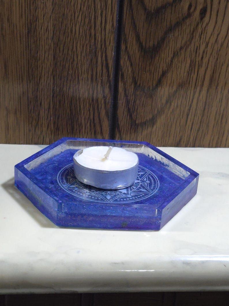Pode incluir: Uma bandeja hexagonal de resina azul com um design circular central apresentando padr&otilde;es geom&eacute;tricos intrincados e motivos de estrelas. Uma pequena vela branca redonda com um inv&oacute;lucro de metal prateado est&aacute; posicionada no centro.