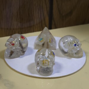 May include: A collection of three clear resin skulls and one clear resin pyramid, each containing embedded white floral elements with small red, blue, yellow, and purple accents. The four objects are arranged on a circular white base.
