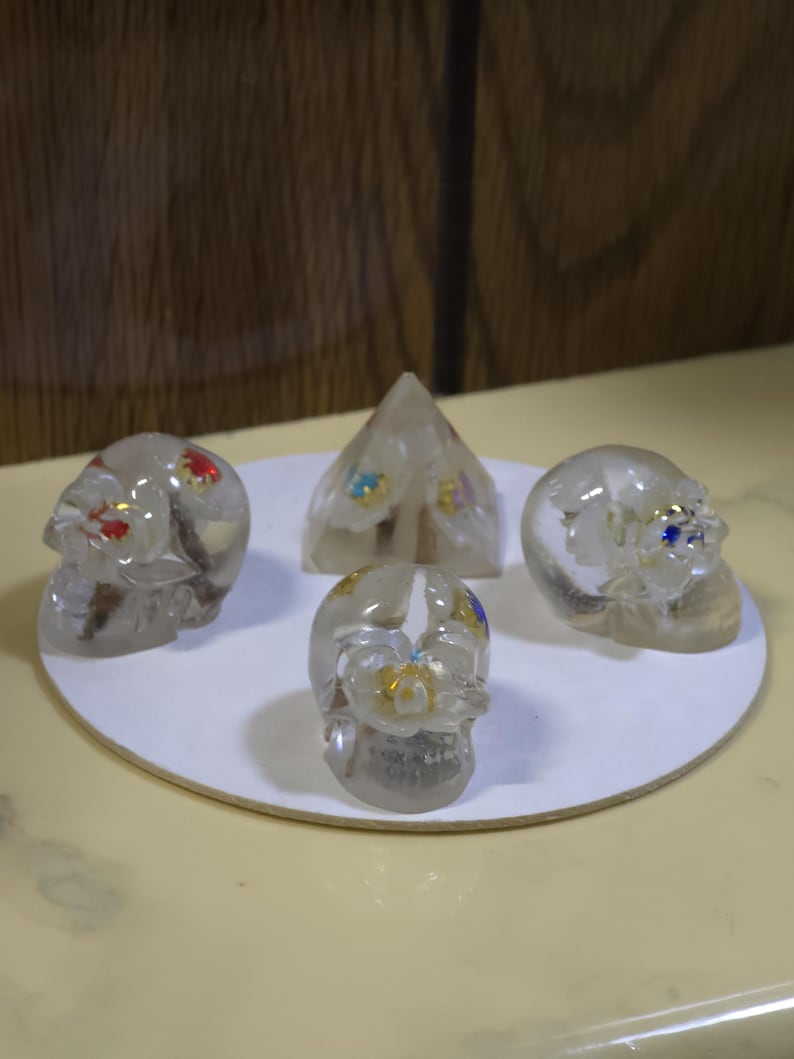 May include: A collection of three clear resin skulls and one clear resin pyramid, each containing embedded white floral elements with small red, blue, yellow, and purple accents. The four objects are arranged on a circular white base.