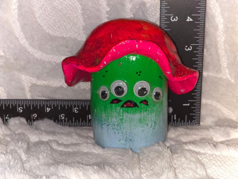 May include: A cylindrical green and blue creature figurine with a wide red mushroom-like cap, four googly eyes, and a painted mouth. A black ruler is positioned behind and to the side, showing a height of approximately 3.5 inches.