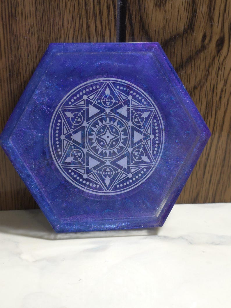Pode incluir: Um porta-copos hexagonal com acabamento em resina brilhante em azul e roxo profundo. O centro apresenta um design geom&eacute;trico de mandala branco e intrincado, composto por tri&acirc;ngulos, c&iacute;rculos e padr&otilde;es em forma de estrela, criando um efeito visual detalhado, sim&eacute;trico e em camadas.
