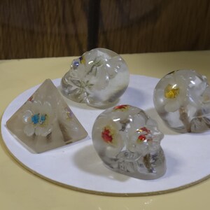 May include: Three clear resin skulls and one clear resin pyramid, each containing embedded dried flowers. The skulls feature white, yellow, and red floral accents, while the pyramid contains white and blue flowers. These decorative objects are arranged together.