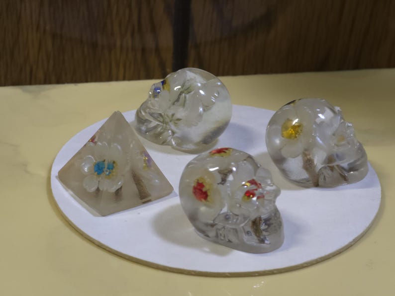 May include: Three clear resin skulls and one clear resin pyramid, each containing embedded dried flowers. The skulls feature white, yellow, and red floral accents, while the pyramid contains white and blue flowers. These decorative objects are arranged together.