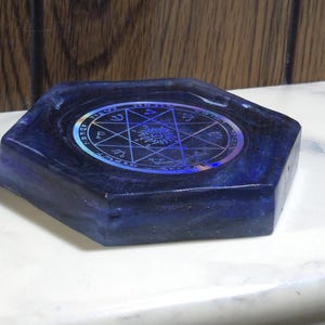 May include: A hexagonal dark blue resin tray featuring a central circular holographic inlay. The inlay displays a complex geometric star pattern, a sun symbol, and various astrological or occult-style symbols arranged in a circular border.