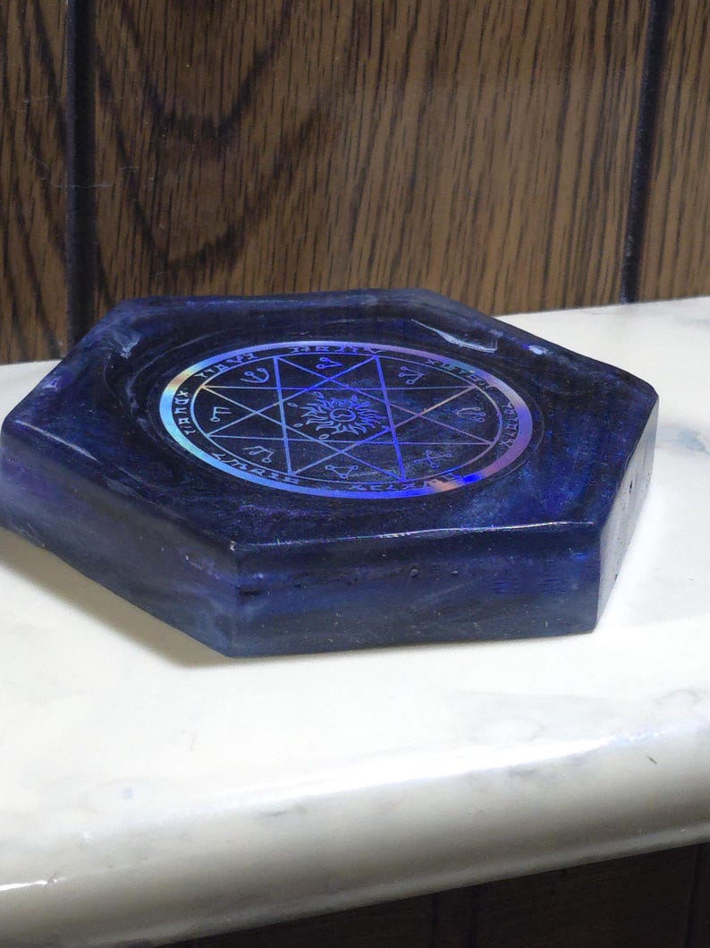 May include: A hexagonal dark blue resin tray featuring a central circular holographic inlay. The inlay displays a complex geometric star pattern, a sun symbol, and various astrological or occult-style symbols arranged in a circular border.