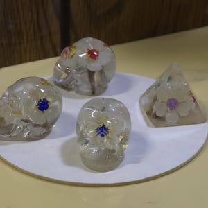 May include: Two clear resin skulls and one clear resin pyramid, each containing embedded white flowers with small colorful centers. The skulls feature floral details on the sides and top, while the pyramid displays a single flower on its front face.