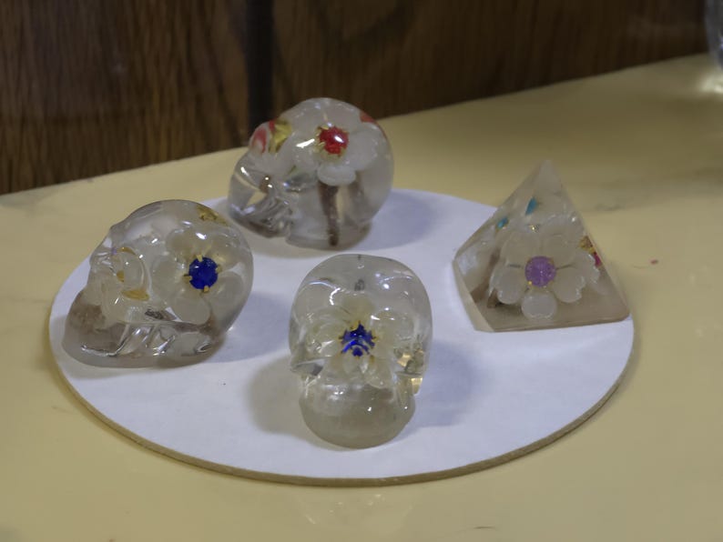 May include: Two clear resin skulls and one clear resin pyramid, each containing embedded white flowers with small colorful centers. The skulls feature floral details on the sides and top, while the pyramid displays a single flower on its front face.