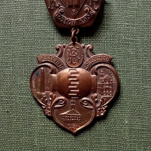 May include: A vintage bronze-colored convention pin. The top section features a pot-shaped design with the text "K.F.C.B. BOSTON 1903". The bottom section has a shield shape with the text "TWENTY FIFTH CONVENTION" and illustrations of buildings.