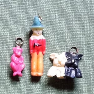 May include: Three small, colourful charms. One is a pink monkey, another is a figure in a blue hat and red coat, and the last is a pair of dogs, one black and one white. Each charm has a silver-coloured loop.