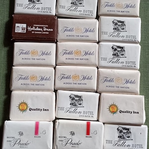 May include: A selection of vintage hotel soaps and a matchbook. The soaps are white, featuring names like "The Fallon Hotel," "Fields Hotels," "Quality Inn," and "Hotel Del Prado." The matchbook is brown and says "Holiday Inns" of Canada Limited.
