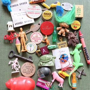 Peut inclure: Une collection de bibelots et petits jouets vintage comprenant une poupée troll aux cheveux verts, un cochon en plastique rouge, une clé en métal, un lézard vert, divers bouchons, capsules de lait, figurines en plastique et jetons publicitaires.