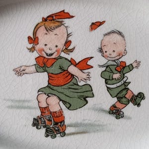 May include: A vintage ceramic plate featuring a whimsical illustration of two children roller skating. The children are dressed in green and orange outfits, with red bows in their hair. The plate has a white background with a red border.