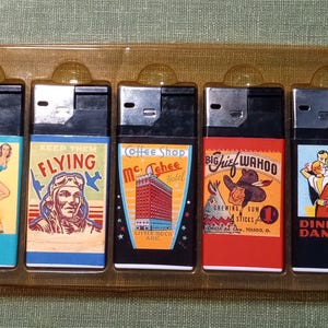 Może przedstawiać: Zestaw pięciu zapalniczek w stylu vintage w przezroczystym plastikowym etui. Każda zapalniczka ma inny, kolorowy wzór graficzny. Wzory obejmują kobietę z psem, pilota, hotel, rdzenną Amerykankę i tańczącą parę. Zapalniczki są czarne i srebrne.