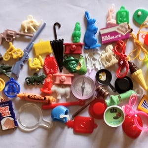 Może przedstawiać: Kolekcja małych, kolorowych plastikowych zawieszek i drobiazgów. Zawiera miniaturowy pistolet, motocykl, niebieskiego królika, kompas, kredkę, lupę i baton czekoladowy. Słowo "Crayon" jest widoczne na jednym z przedmiotów.
