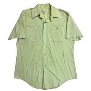 May include: A light green short-sleeved button-down shirt with two chest pockets. The shirt has a pointed collar and a label at the neck. The shirt is made of a lightweight fabric.