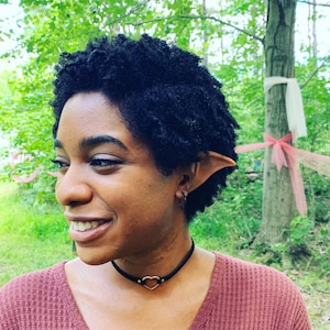 May include: A person with short, curly black hair wearing a maroon sweater and a black choker necklace with a heart pendant. They are wearing a pair of elf ears.
