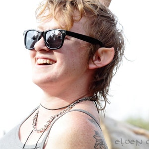 May include: A person with short, light brown hair wearing black sunglasses and a silver chain necklace. The person is smiling and looking to the right.