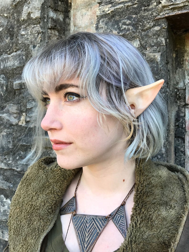 Puede incluir: Una persona con el pelo corto gris plateado que lleva una chaqueta verde peluda y un collar con un dise&ntilde;o geom&eacute;trico. La persona lleva orejas de elfo.