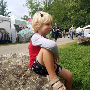 Puede incluir: Un niño pequeño con una camisa roja y pantalones cortos negros se sienta en una roca grande. Lleva un accesorio de ala de mariposa verde y azul en la espalda y una diadema de cuerno de diablo negro. El niño lleva sandalias y mira hacia la derecha.