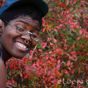 May include: A person with a blue baseball cap and glasses is wearing a silver nose ring and a silver lip ring. They are smiling and looking at the camera. The background is a blurry image of red leaves.