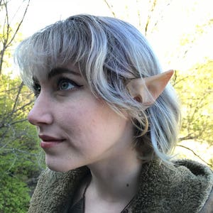 Puede incluir: Una persona con el pelo corto plateado que lleva una chaqueta verde con capucha y un collar marr&oacute;n con un dise&ntilde;o geom&eacute;trico. Lleva orejas de elfo.