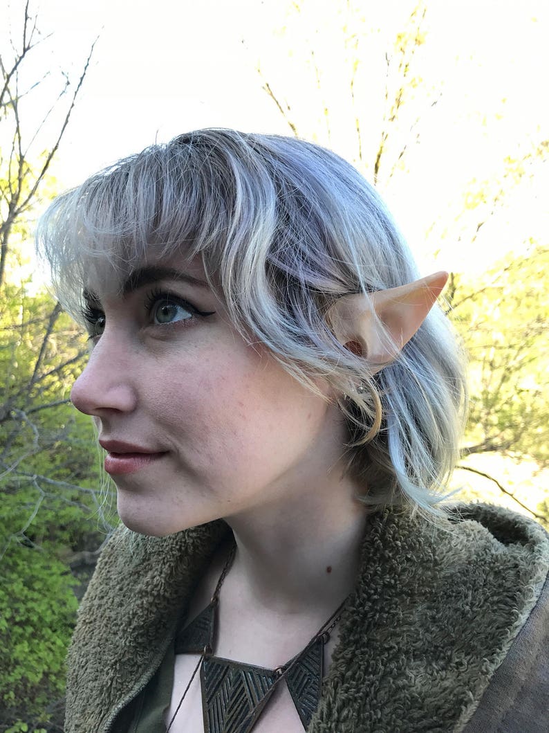 Puede incluir: Una persona con el pelo corto plateado que lleva una chaqueta verde con capucha y un collar marr&oacute;n con un dise&ntilde;o geom&eacute;trico. Lleva orejas de elfo.