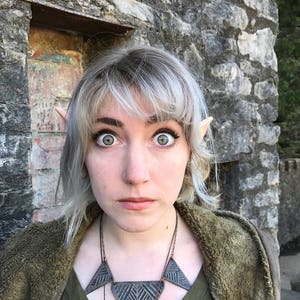 Puede incluir: Una mujer con el pelo corto plateado y orejas puntiagudas lleva una camisa verde y un collar con tres colgantes triangulares. Est&aacute; de pie frente a una pared de piedra.