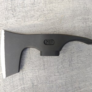 May include: A black axe head with a white edge and a black handle. The axe head has a rounded shape and a small hole in the top. The handle has a black plastic grip and a metal ring at the end. The axe head is stamped with the letters "MF".