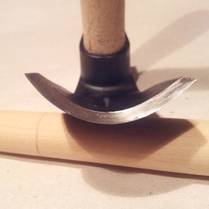 Puede incluir: Un primer plano de una herramienta de talla de madera con una cuchilla curva y un mango de madera. La herramienta está apoyada sobre una pieza de madera.