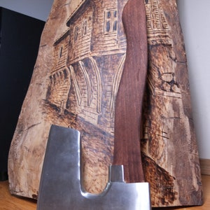 May include: A large, silver axe with a wooden handle. The axe is resting on a piece of wood with a burned image of a building.
